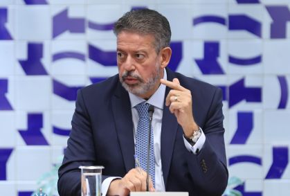 Arthur Lira, sentado, olha para o lado direito enquanto fala e gesticula com a mão esquerda, com o indicador em riste. Na mão direita, segura uma caneta