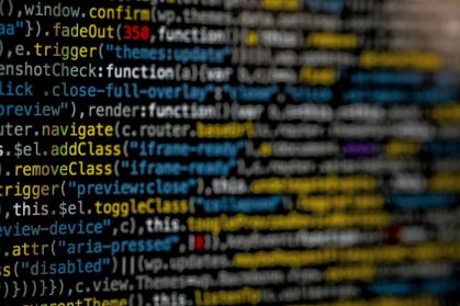 A fotografia apresenta uma tela de computador com uma sequência de códigos de programação, com letras nas cores amarelo, branco, azul, cinza e vermelho.